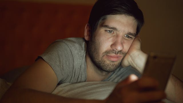 一个男人右手拿手机，左手撑脸，在昏暗的环境下刷手机